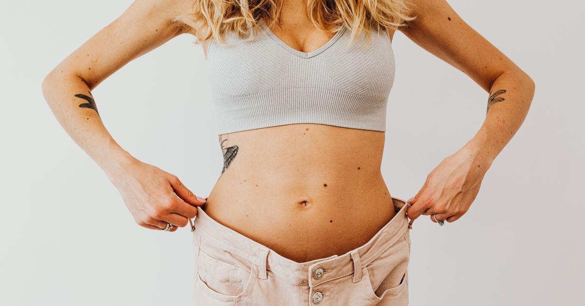 A woman displaying weight loss by wearing oversized pants and crop top, showing tattoos.