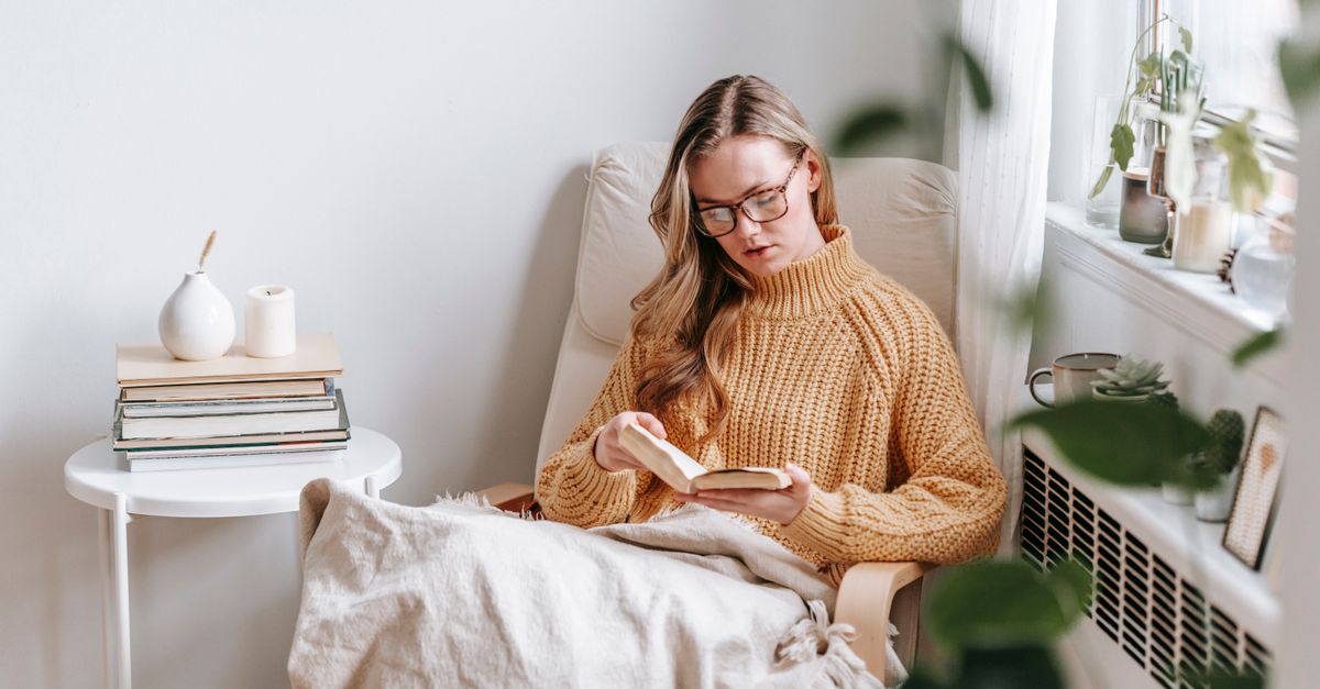 Young female student in trendy sweater and glasses sitting in white armchair with plaid near table with books and candle while reading book