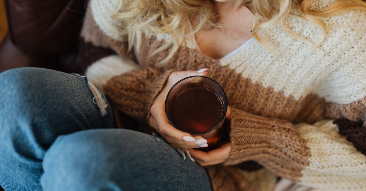 A woman in a cozy sweater and jeans holds a glass of tea, creating a warm indoor moment.