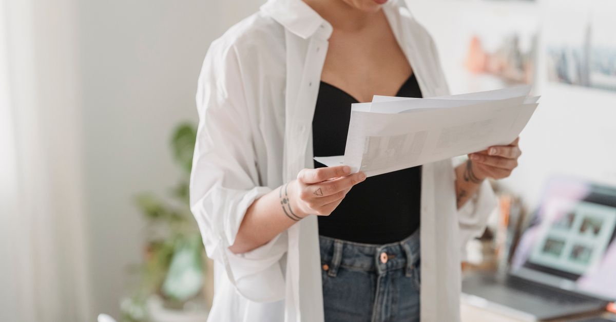 Crop young thoughtful woman with tattoos and paper sheets working against laptop at home in daytime