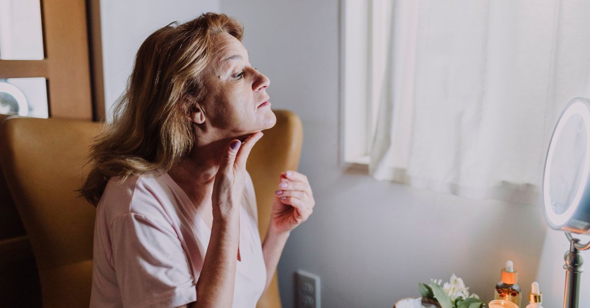 A mature woman examining her skin in a well-lit room with skincare products nearby.