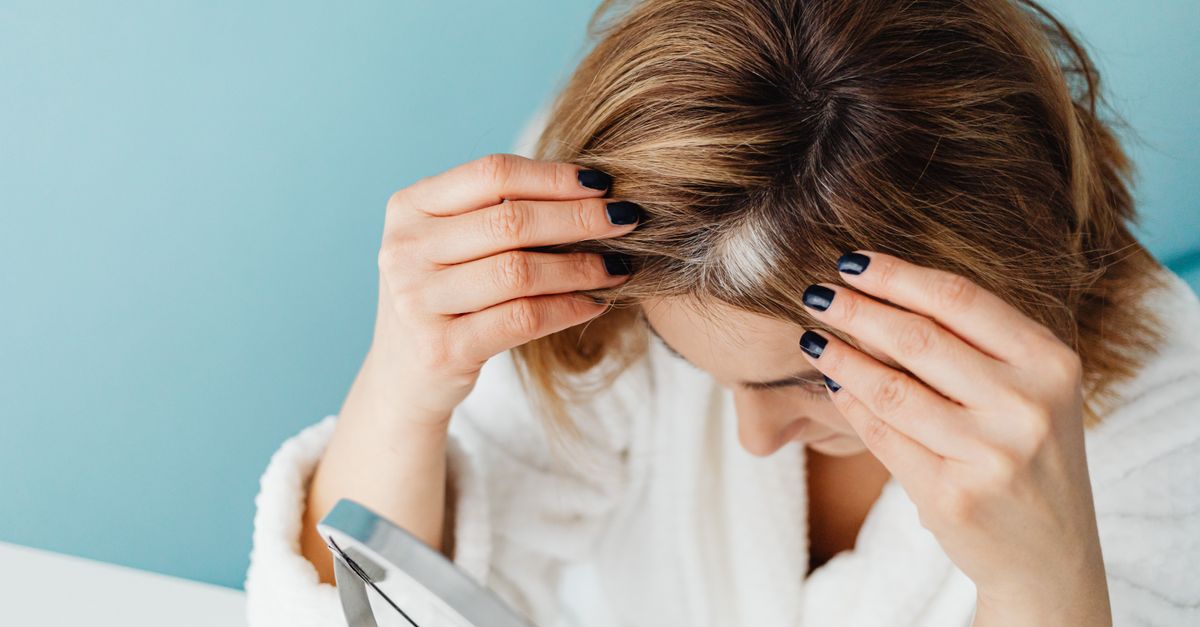 Woman in white robe checking her hair for gray strands using a mirror on blue background.