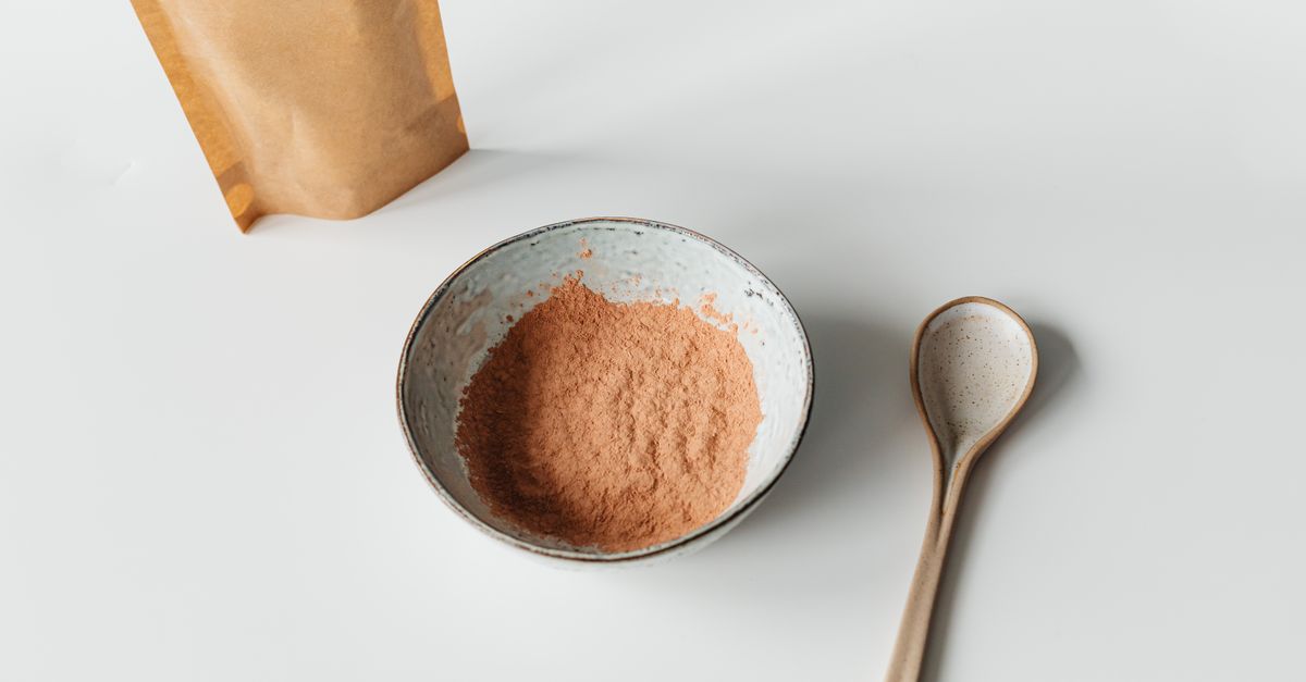 Flat lay of a ceramic bowl with powder, wooden spoon, and paper bag on a white background.