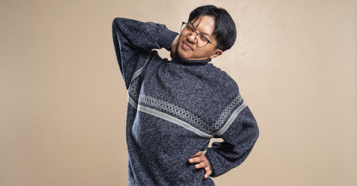 Asian man wearing eyeglasses and sweater stretching indoors, feeling body pain.