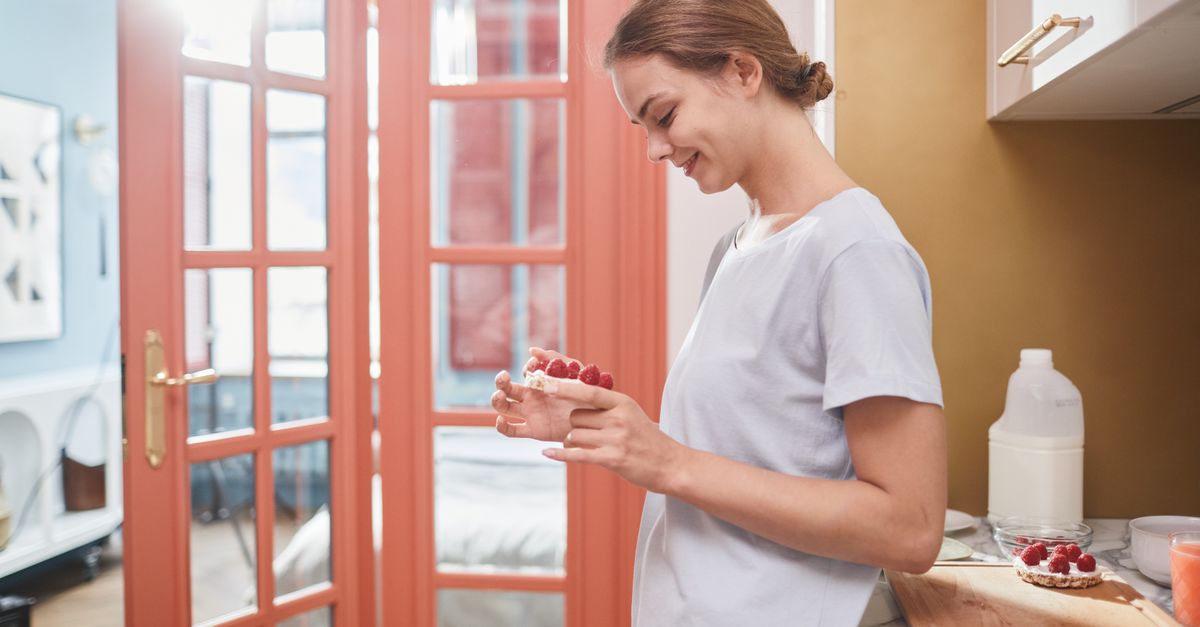 Smiling woman in kitchen holding a healthy breakfast with raspberries, enjoying her morning routine.
