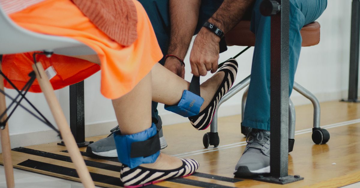 A therapist assists a woman during a rehabilitation session involving ankle weights.
