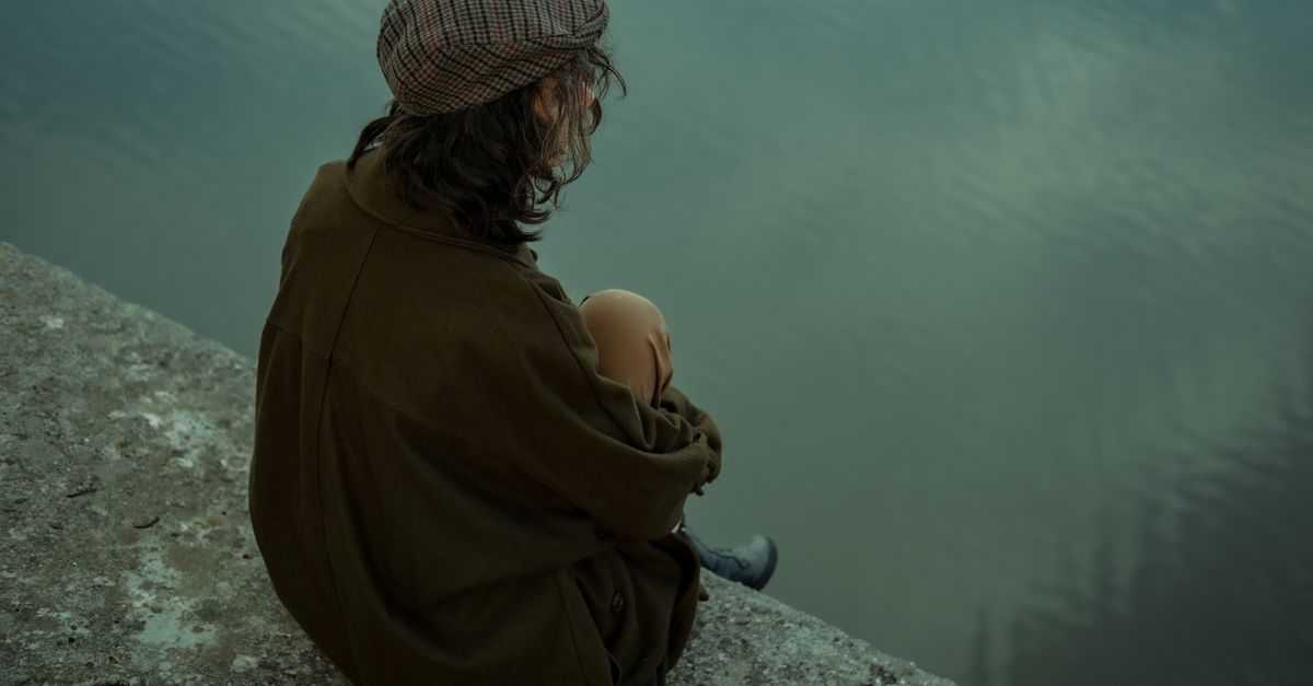 A woman in casual attire sits thoughtfully by the lake, evoking calm and introspection.