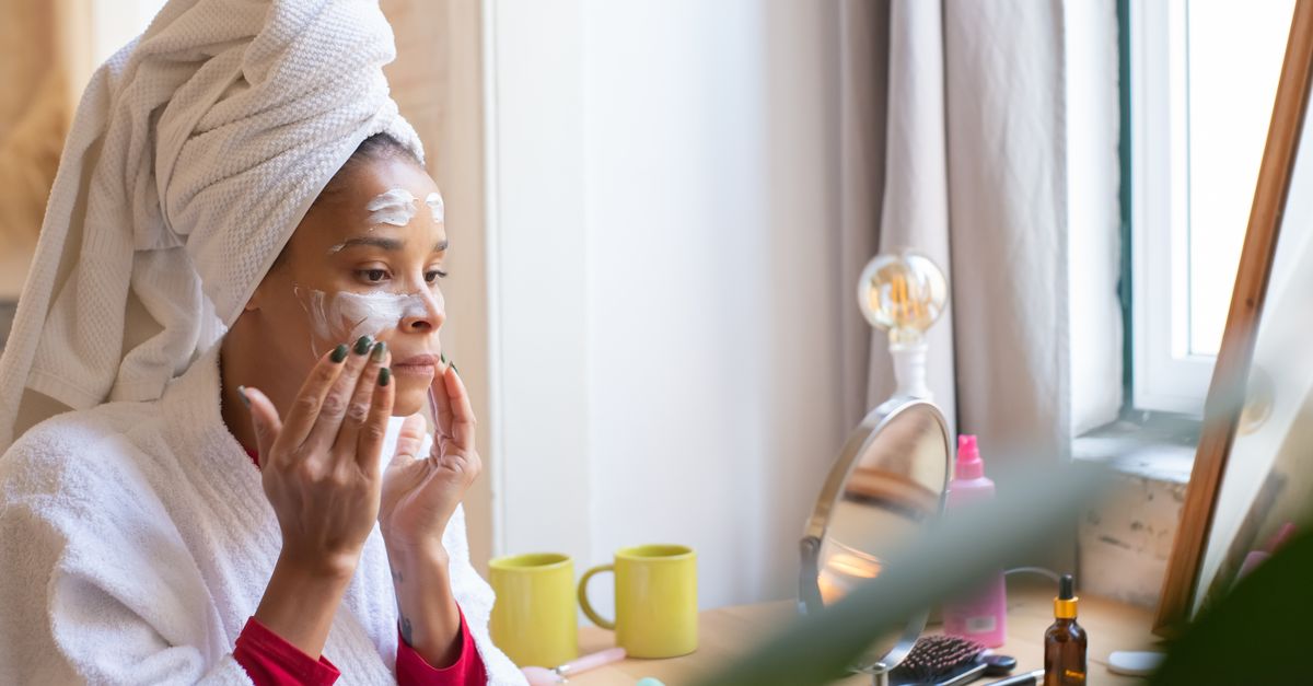 A woman applies face cream in a relaxing home setting, dressed in a bathrobe and head towel.