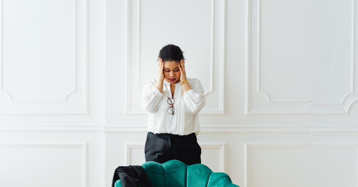 A woman holds her head in stress, standing indoors next to a green chair.