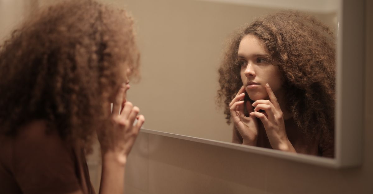 Side view of serious young lady with curly hair in casual clothes exploring face skin while looking at mirror in bathroom
