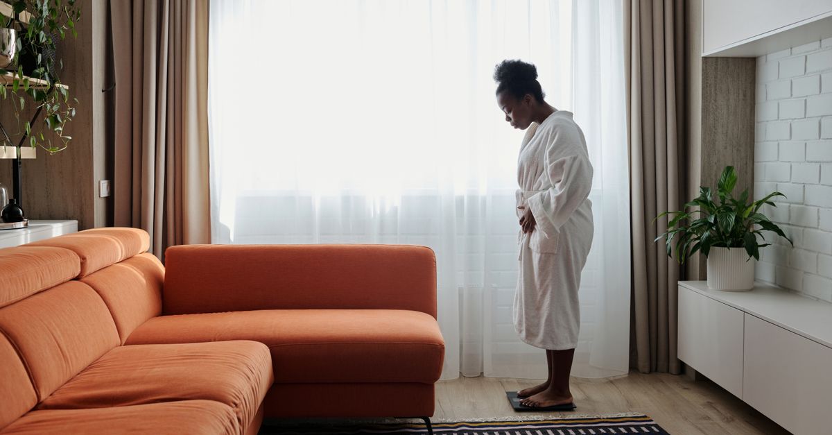 An African American woman in a bathrobe checks her weight on a scale near an orange sofa.