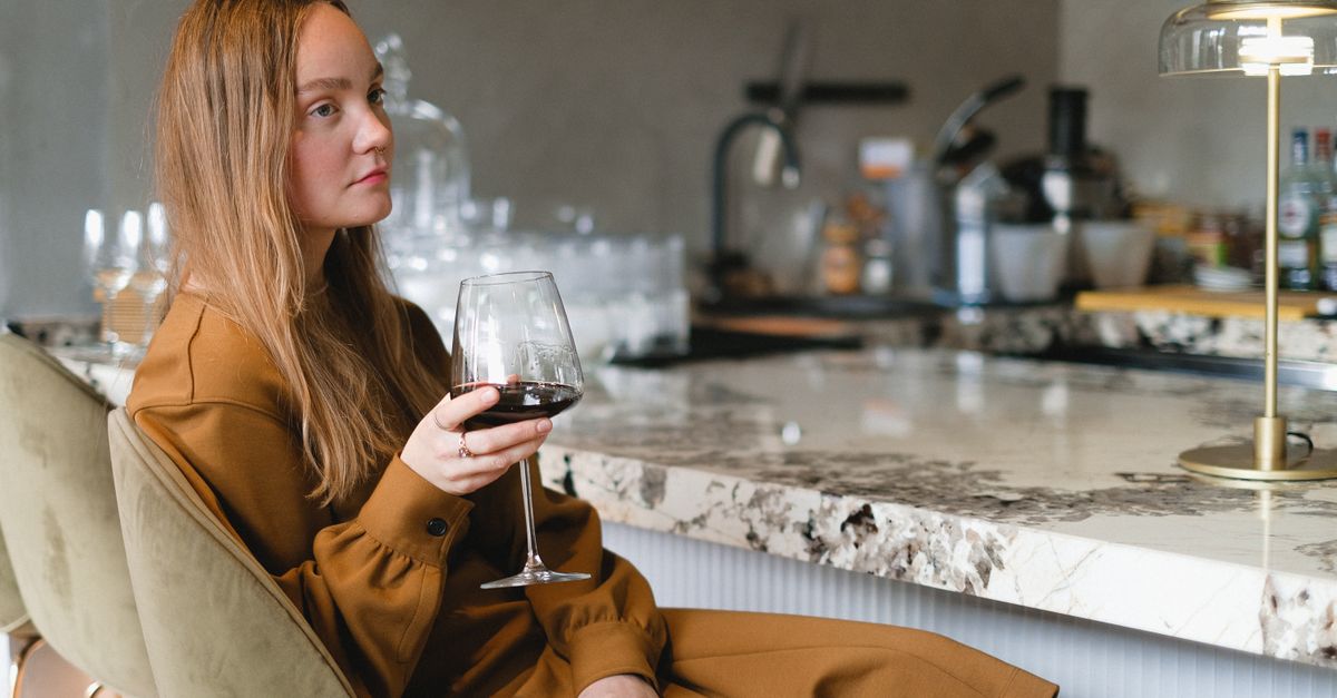 A contemplative woman in a brown dress holding a wine glass at a marble counter.