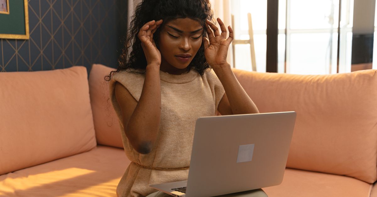 A young woman holding her head in pain while working on a laptop in a cozy home interior.
