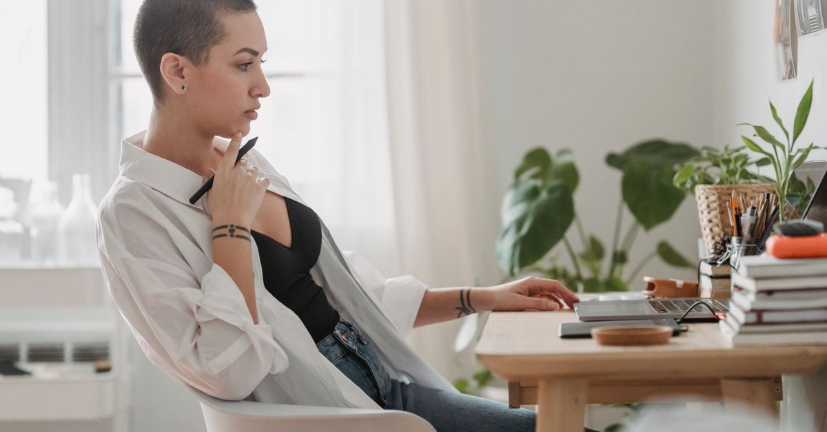 Side view pondering young female wearing casual outfit browsing netbook and touching chin in thoughts while sitting at desk in light room