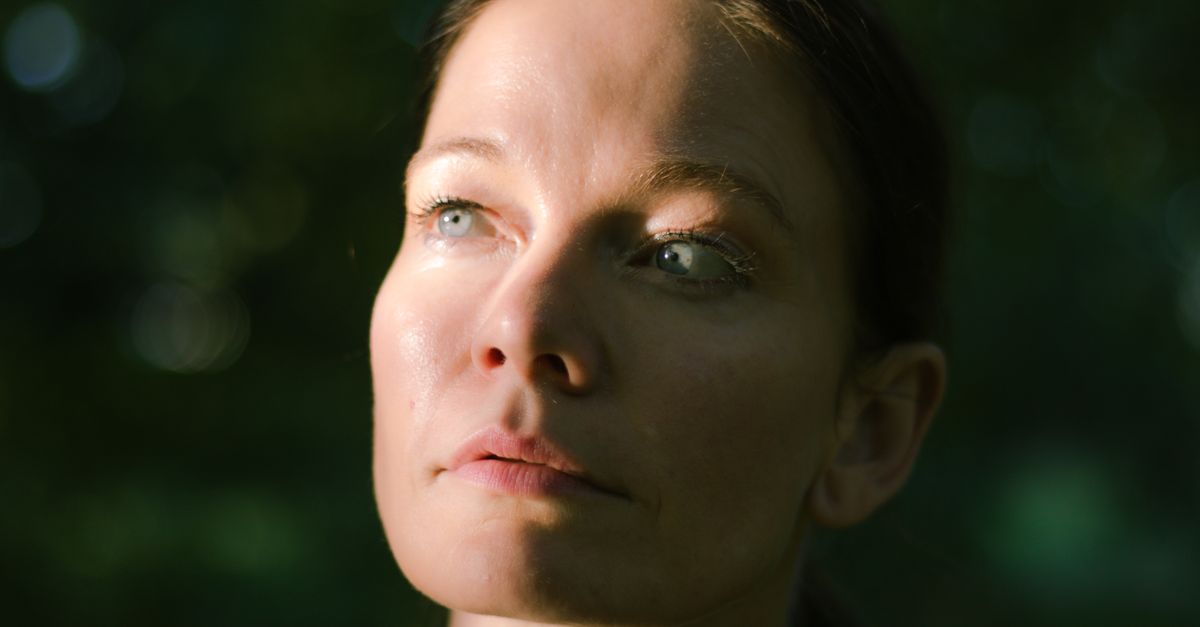 A contemplative woman looks upward in a serene outdoor setting with soft natural lighting.