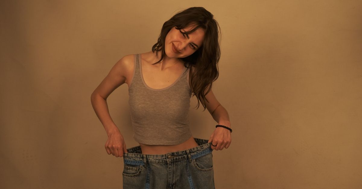 A woman smiles while holding out the waistband of oversized jeans to show her weight loss success.