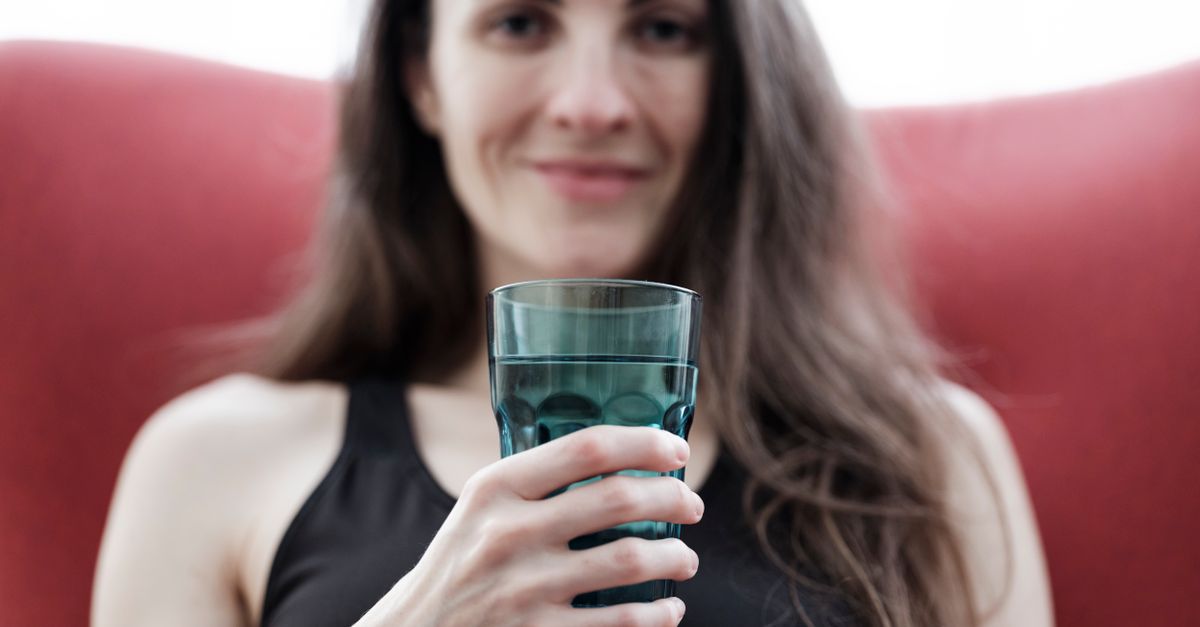 Smiling woman with long hair holding a glass of water, seated indoors with a red chair.
