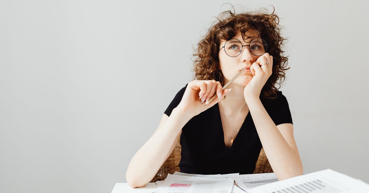 Curly-haired woman with glasses pensively thinking at a desk with documents.