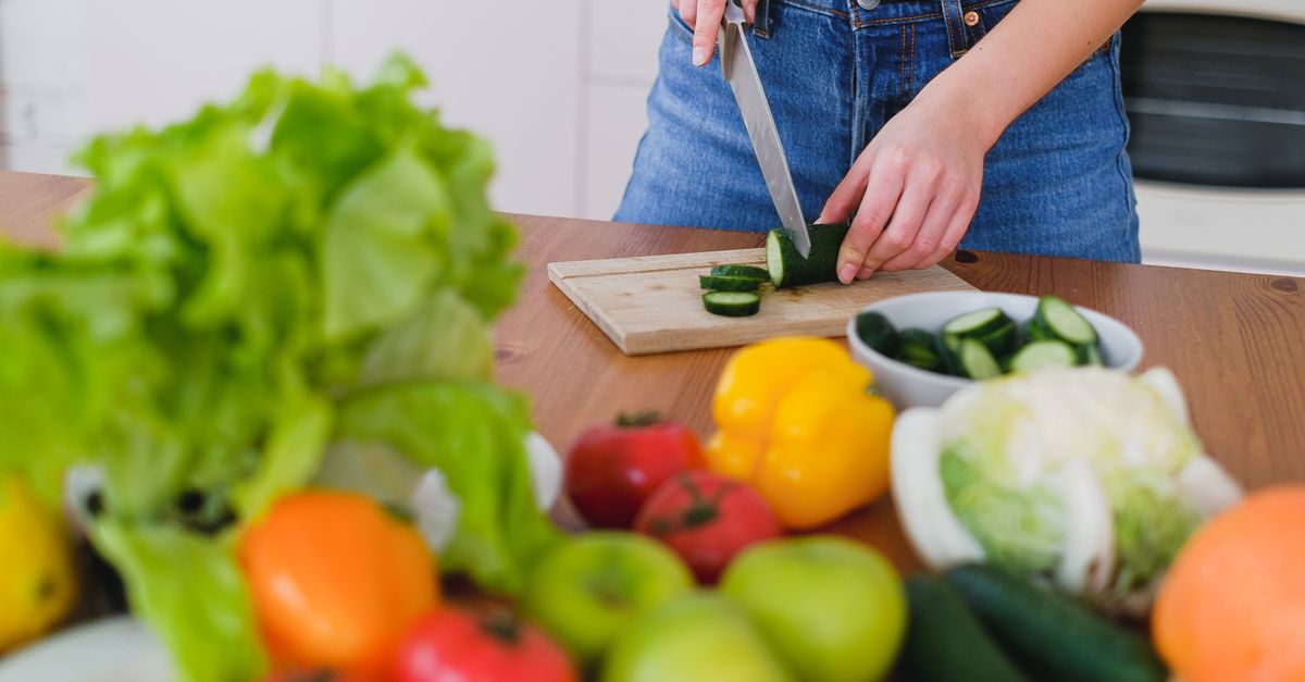 Woman slicing cucumbers surrounded by fresh produce in a bright kitchen setting.