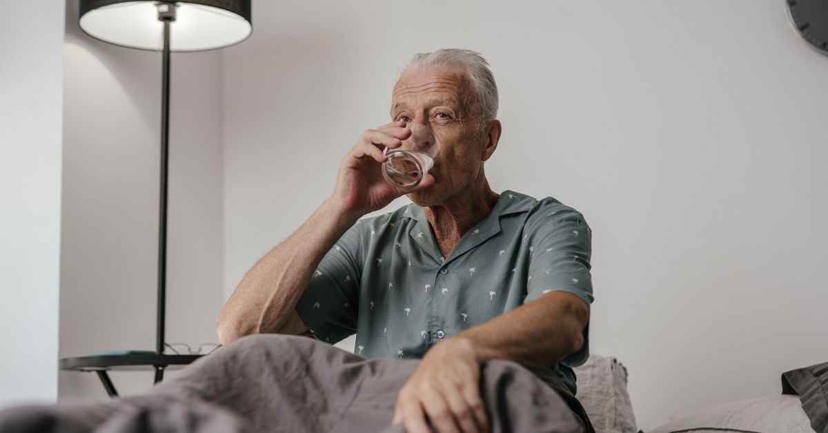 Senior man with gray hair drinks water in bedroom setting, conveying calm and routine.