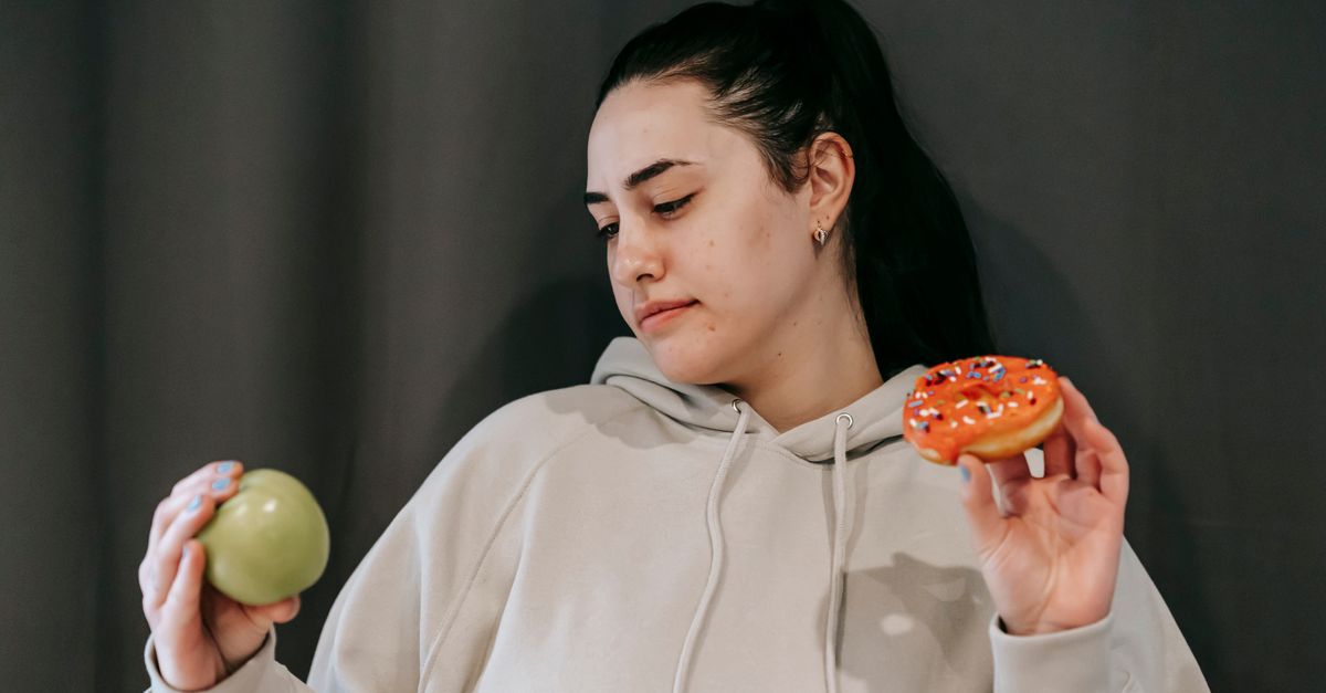 Pensive female wearing casual hoodie deciding between healthy green apple and sweet doughnut in studio