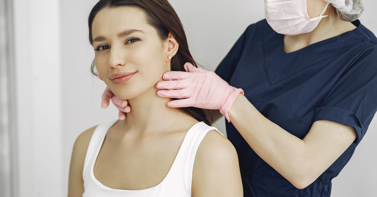 A medical practitioner in scrub suit performs a neck examination on a smiling woman in a clinic.