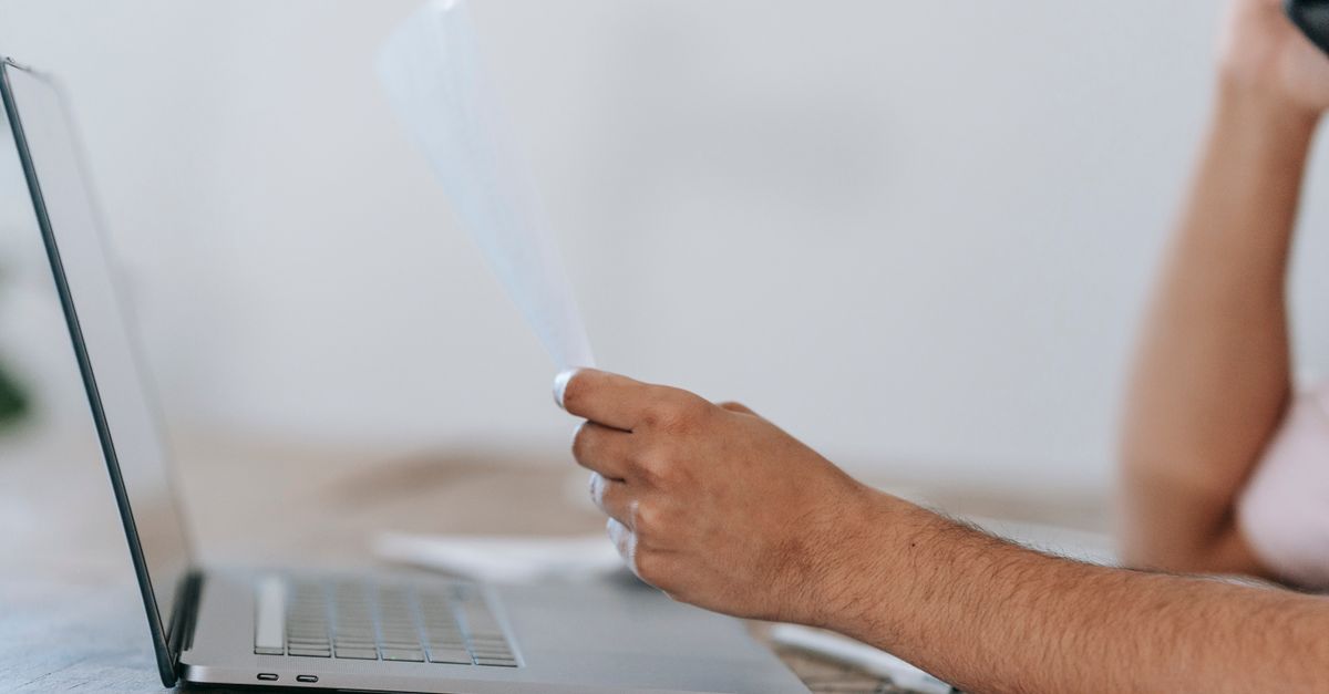 Crop anonymous male reading document and talking on mobile phone while sitting at desk with laptop