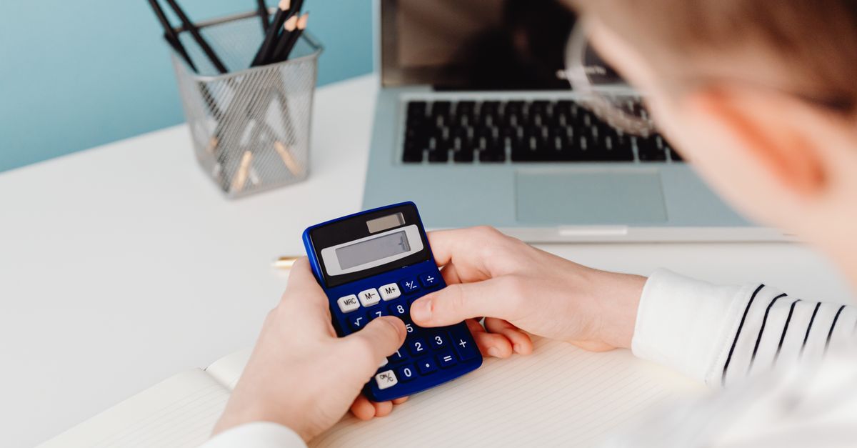 Close-up of hands using a blue calculator at a desk with a laptop and stationery.