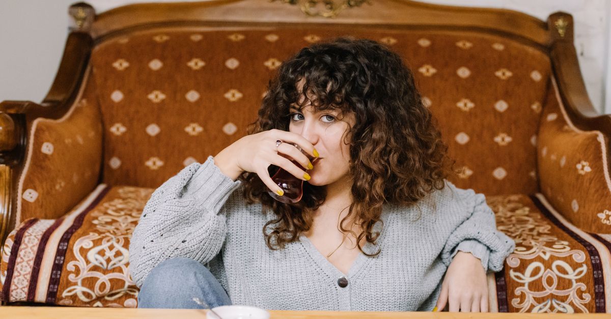 A woman with curly hair relaxing on a vintage sofa, sipping tea indoors.