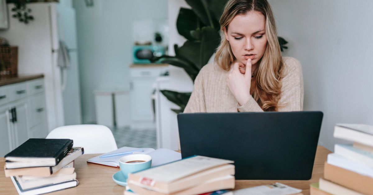 Serious intelligent female studying online on netbook at table with cup of coffee and textbooks on blurred background of home