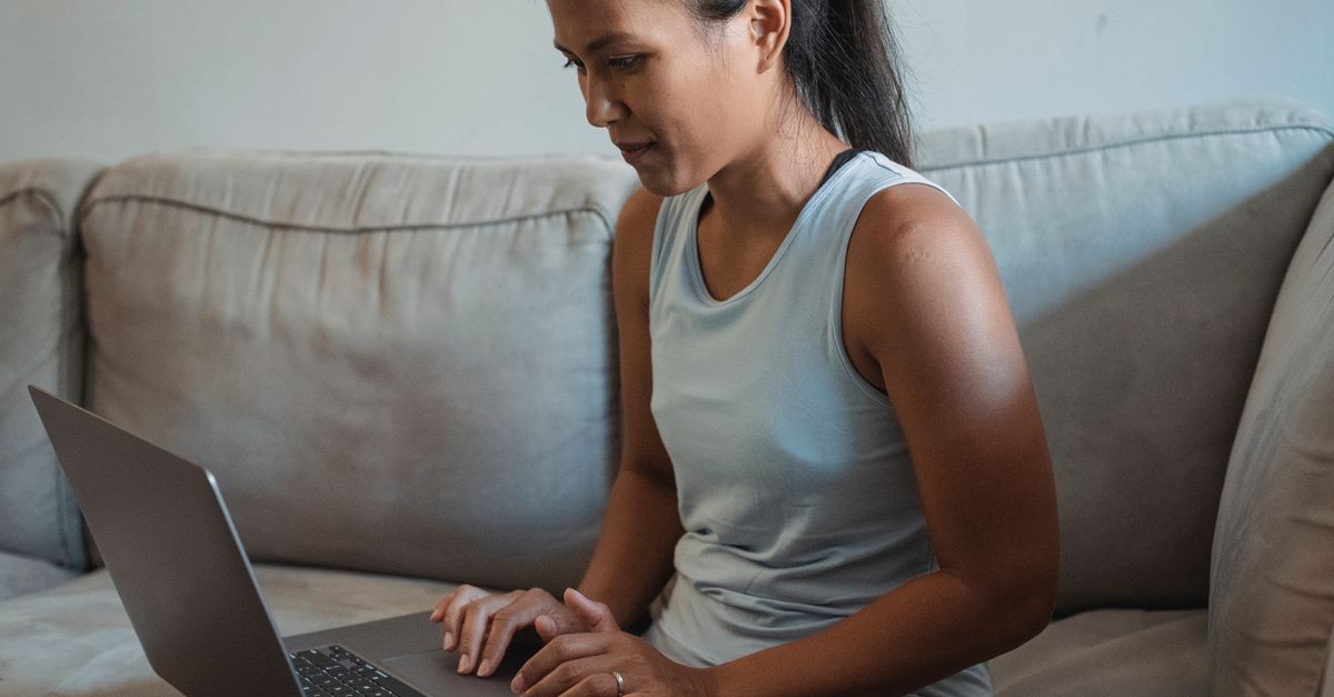 Adult lady sitting on sofa and using computer for remote work in living room while wearing casual outfit