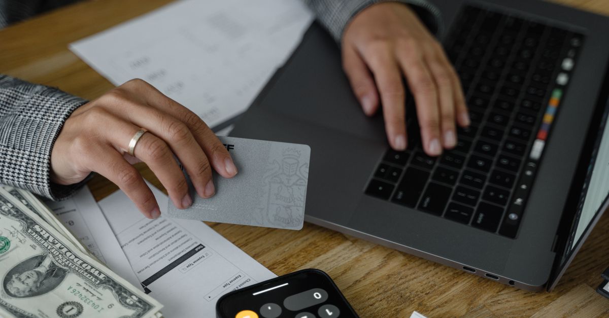 Close-up of a person using a credit card and laptop in a financial setting with cash and calculator.
