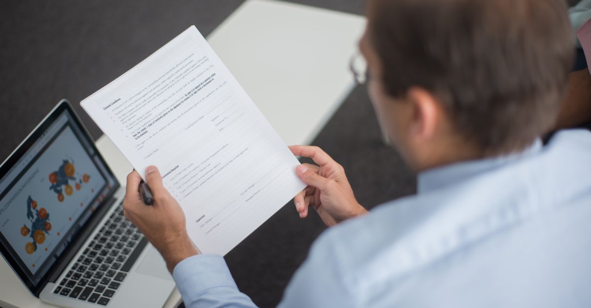 Professional in blue shirt reviewing documents next to laptop displaying charts.