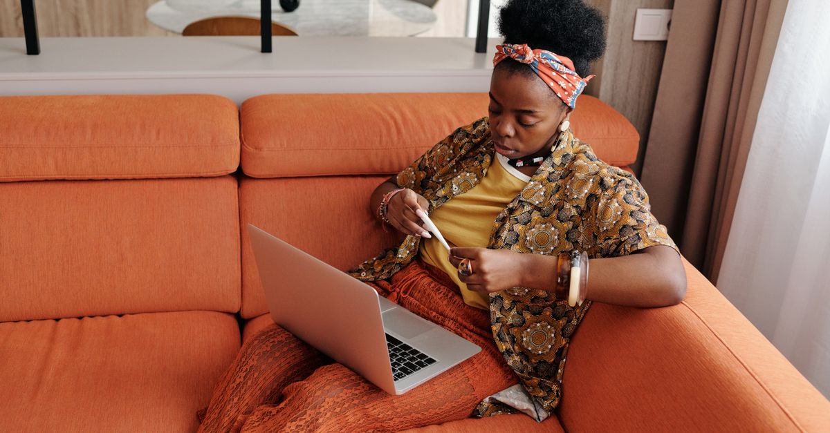 A worried woman sitting on an orange sofa checking her temperature with a thermometer, while a laptop sits open beside her.